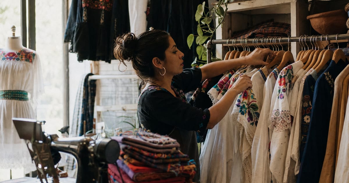 Una mujer joven organiza blusas bordadas en una boutique de ropa, con una máquina de coser vintage y telas dobladas en primer plano.