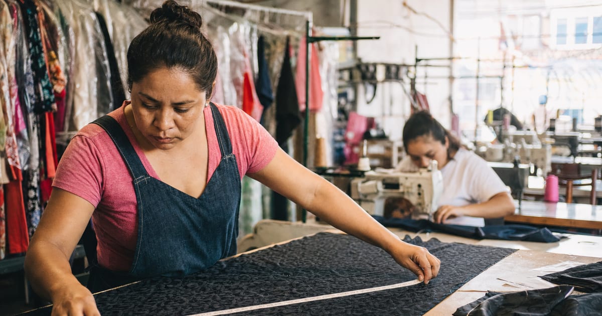 Una costurera mide tela con una cinta métrica en un taller de ropa mexicano, rodeada de prendas y máquinas de coser.
