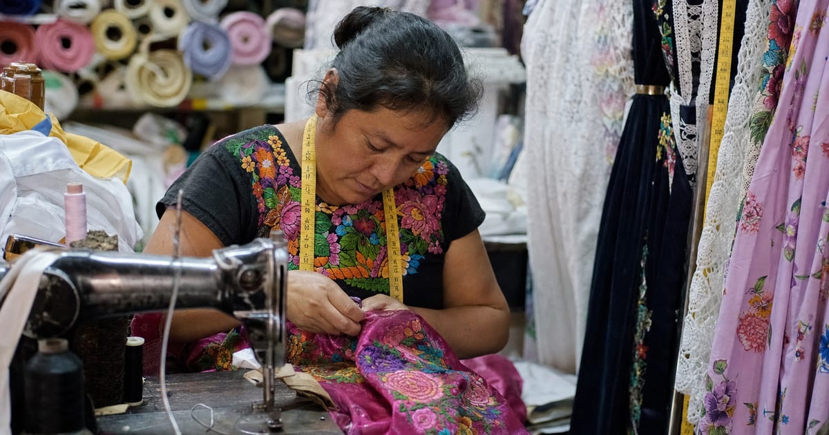 Sastre trabajando en un vestido bordado en un taller de la Ciudad de México, con una cinta métrica colgando cerca.