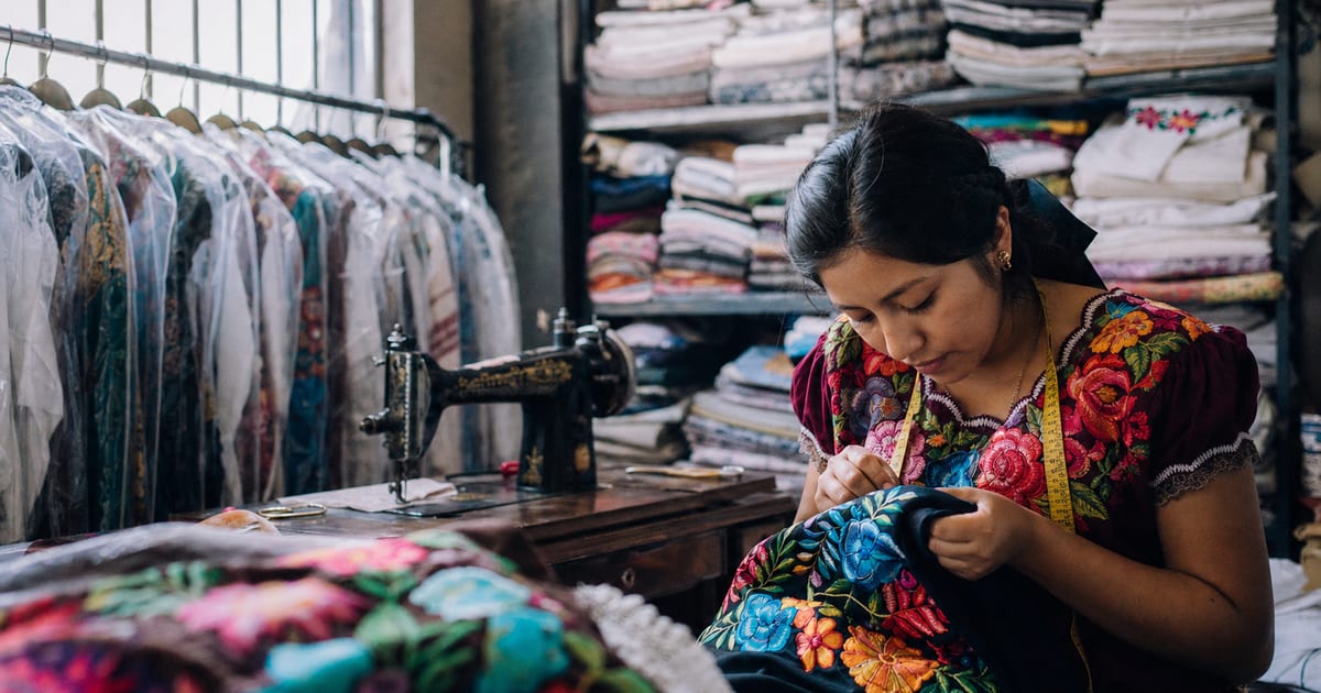 Una costurera joven cose a mano una blusa bordada en un taller de ropa mexicano, con telas y prendas en el fondo.