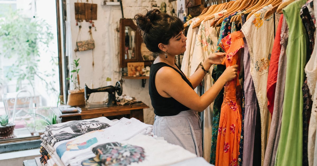 Una mujer joven arregla vestidos en un estante dentro de una boutique de ropa en la Ciudad de México, con una pila de camisetas dobladas en primer plano.