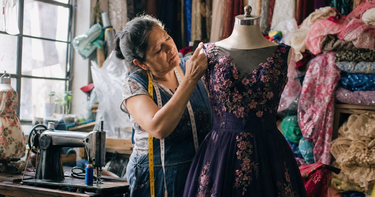 Una costurera ajusta un vestido en un maniquí en un taller de costura mexicano, con telas y una máquina de coser de fondo.