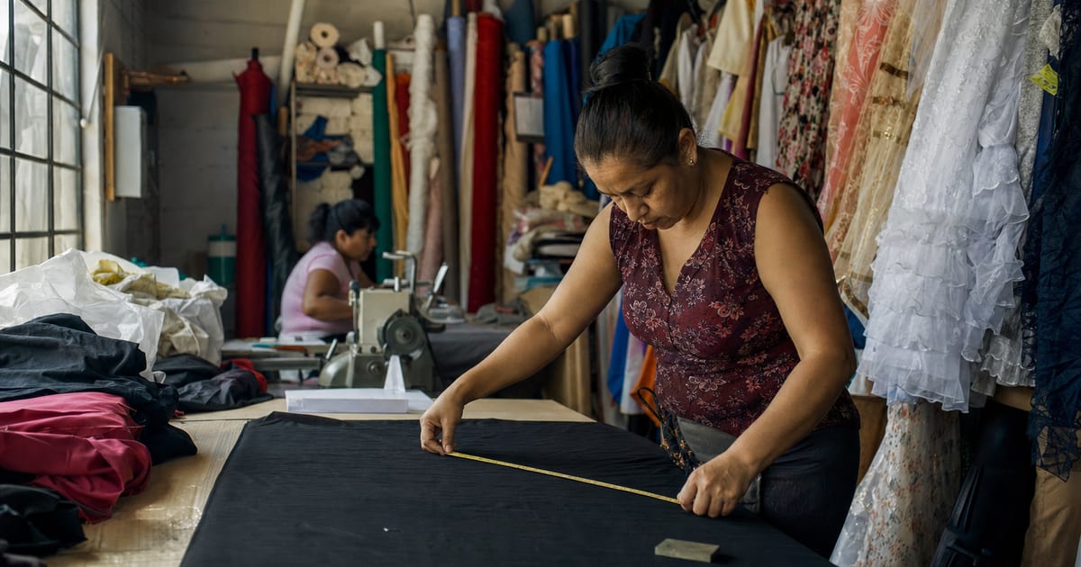 Una costurera mide tela con una cinta métrica en un taller de ropa mexicano, con prendas y rollos de tela alrededor.