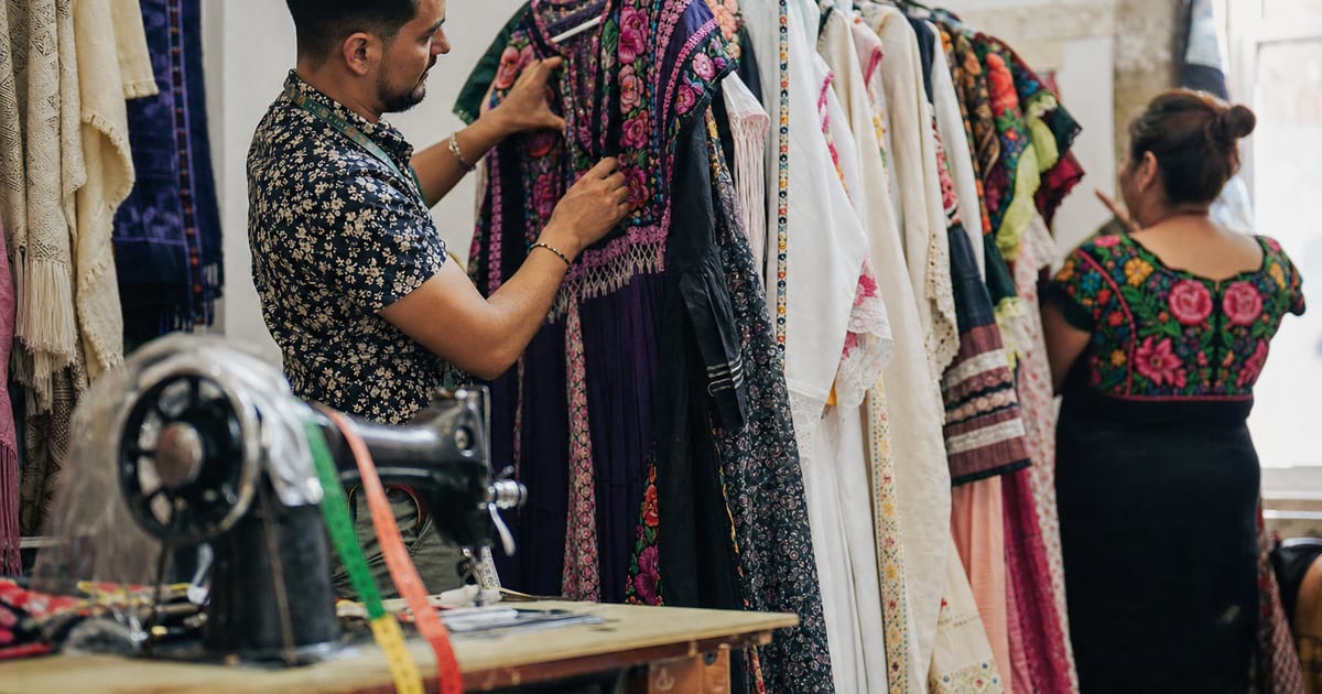 Un sastre o asistente de tienda en una boutique de ropa mexicana, organizando vestidos bordados con una cinta métrica sobre una máquina de coser antigua.