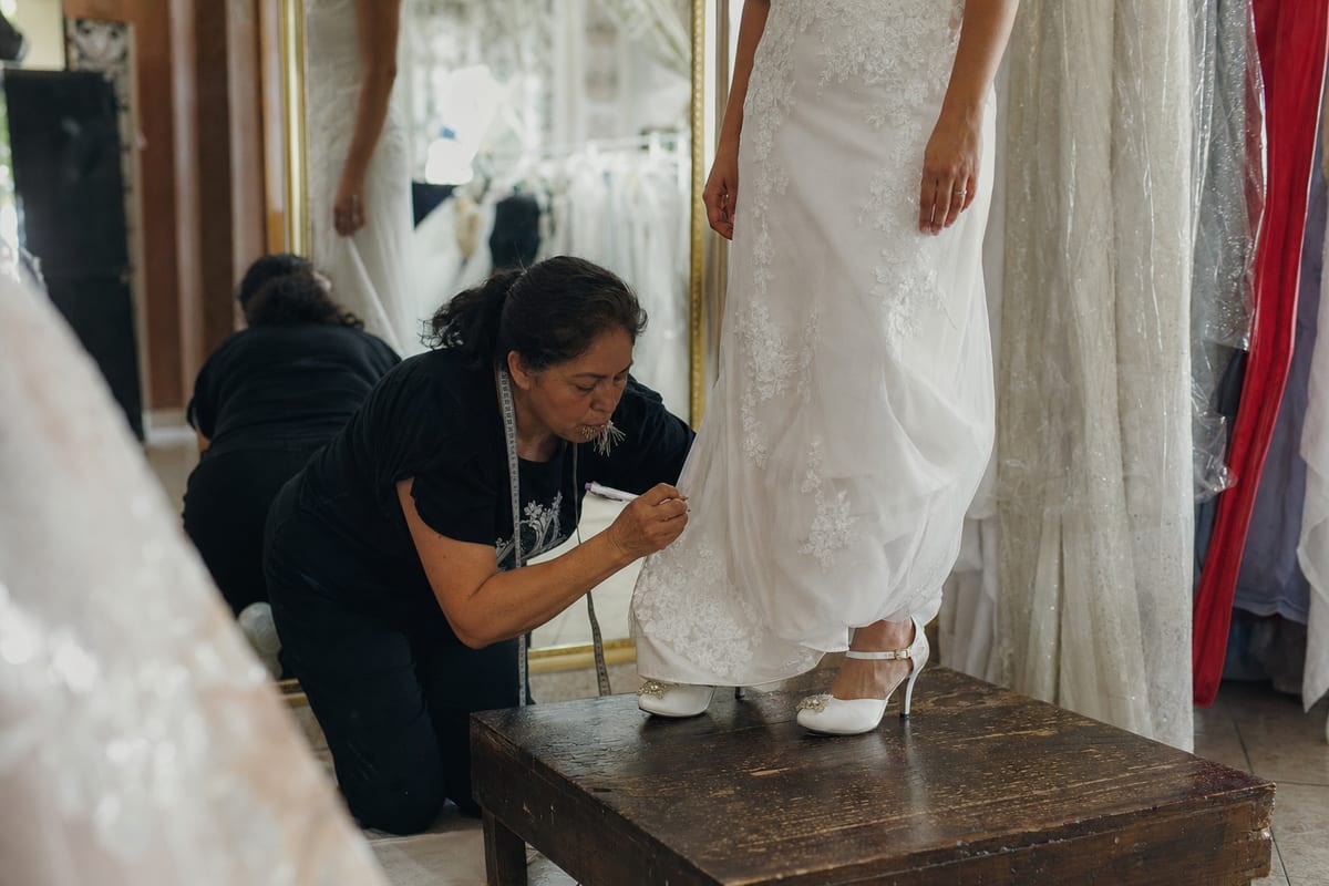 Costurera marcando con alfileres la bastilla de un vestido de novia mientras la clienta está parada sobre una tarima con sus zapatos de la boda